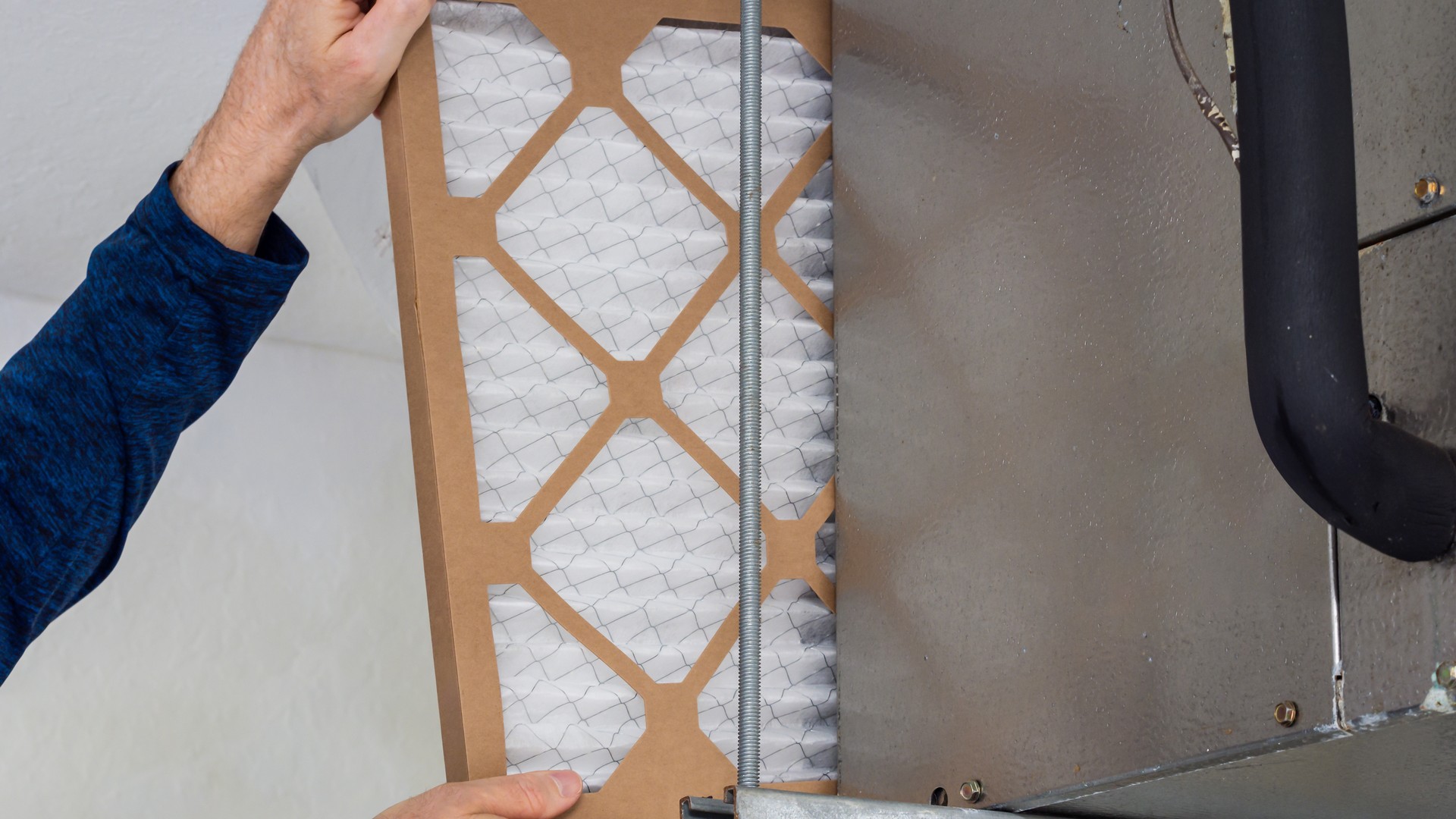 A homeowner inserting a clean white pleated air filter into the filter slot of a residential ducted air handler, replacing a dirty one
