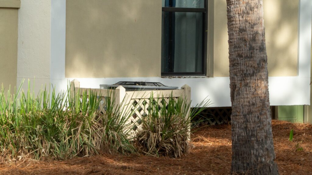 Residential outdoor central air conditioning condenser unit beside a single-family Florida home with a tropical palm tree, green foliage, and scattered pine needles around the base, illustrating the system sitting through a multi-day Treasure Coast storm power outage.