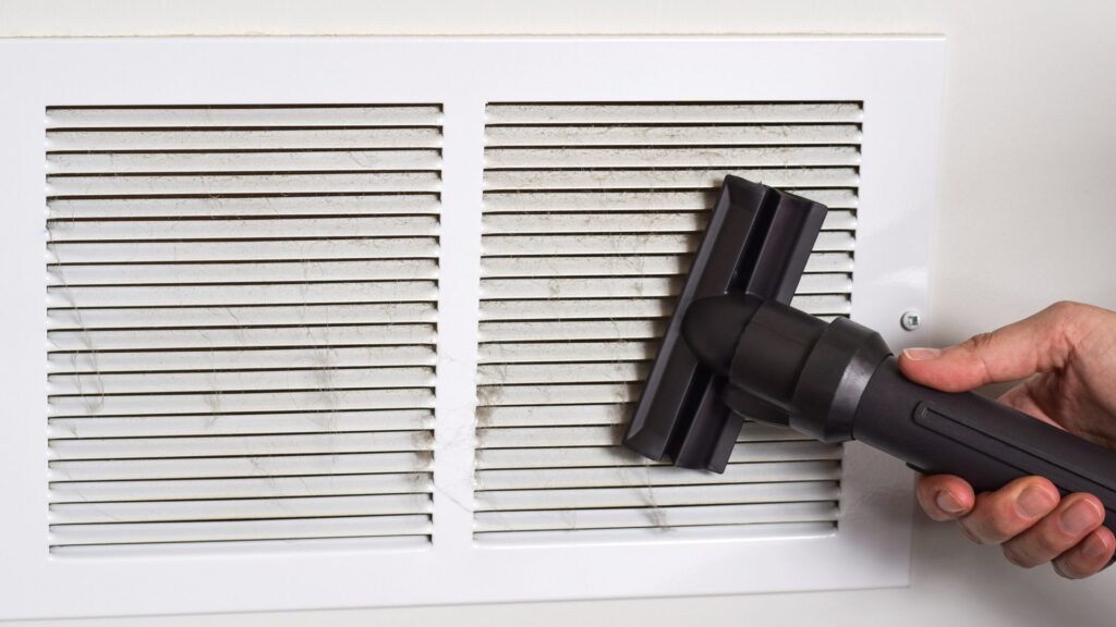 A hand vacuuming dust from a white return-air grille on the wall of a Florida home