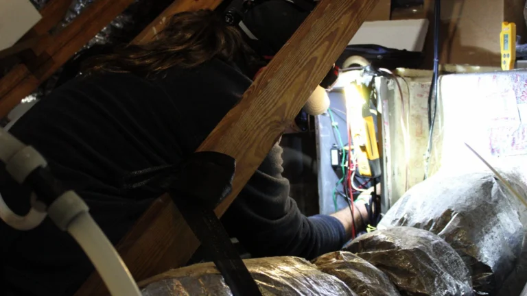 A technician in a dark cap and sweatshirt leans between attic rafters to work at an air handler's control wiring with a yellow meter beside it, with silver-wrapped flex ducts in the foreground and a white pipe at the left.