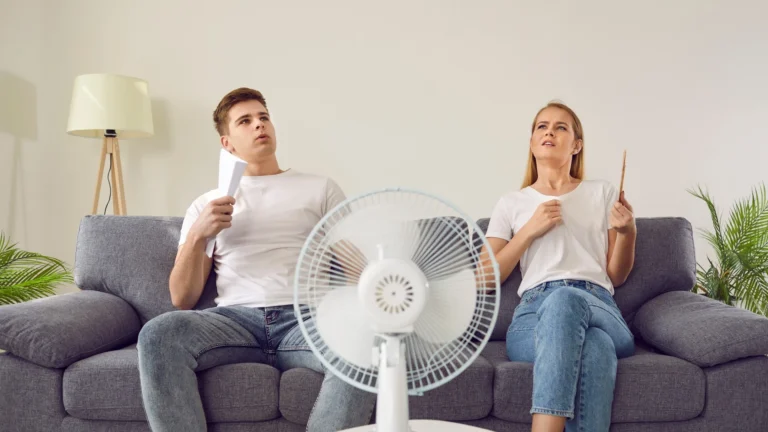 A man and a woman in white T-shirts sit on a gray sofa in a living room, fanning themselves with a sheet of paper and a small hand fan while a white pedestal fan runs in front of them.